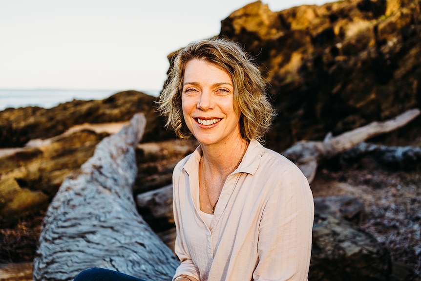 A woman with blonde hair sitting at the beach on a log overlooking the water.