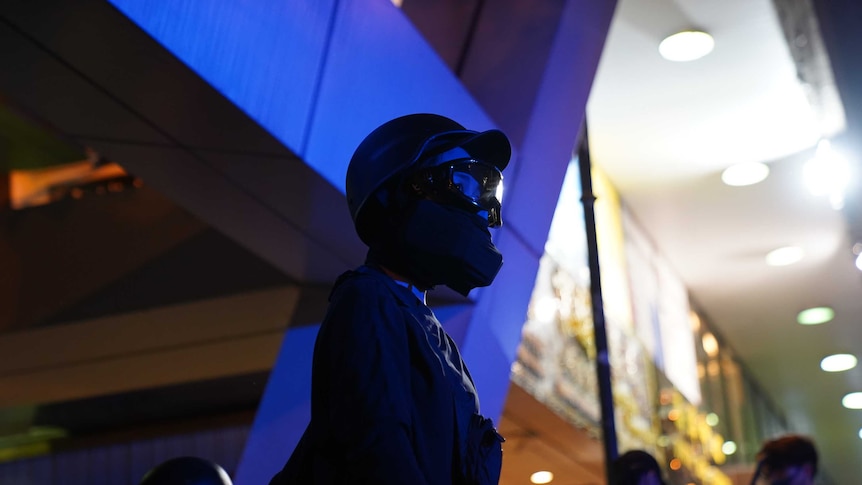 A protester clad in black is wearing a helmet and gas mask standing on front of a blue-lit building.