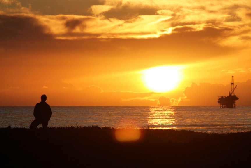 Silhouette of a man standing on a shoreline at sunset looking out at an oil rig on the ocean.