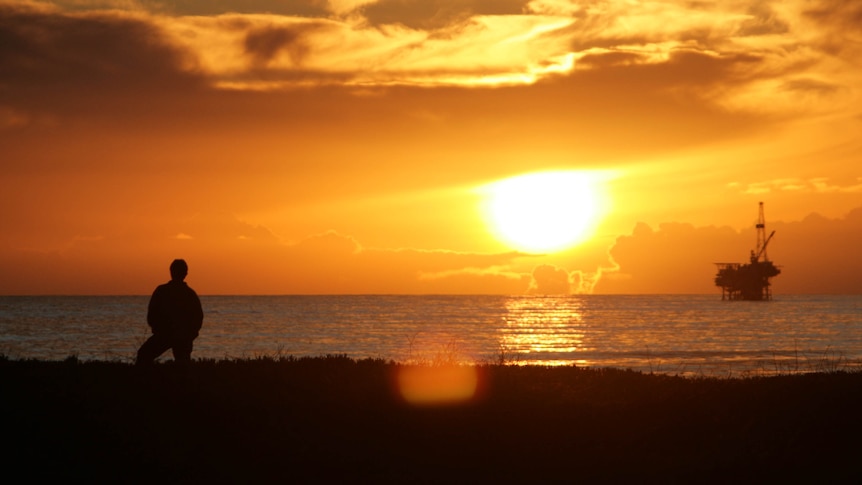 Silhouette of a man standing on a shoreline at sunset looking out at an oil rig on the ocean.