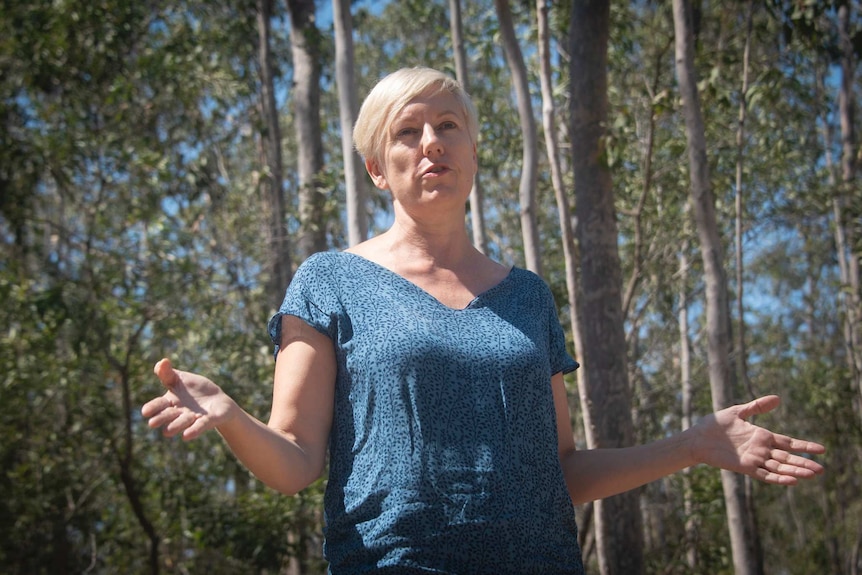 A woman with short blonde hair, wearing a blue top, is speaking.  She is surrounded by trees