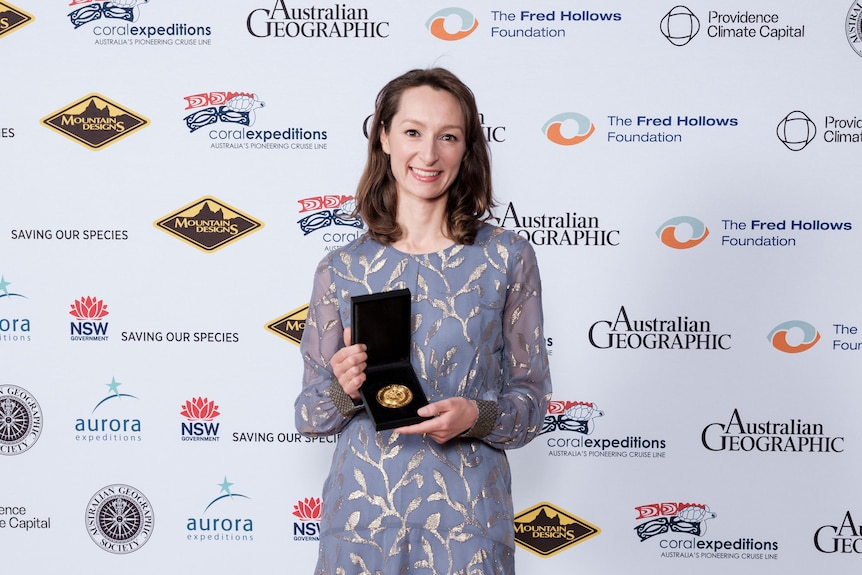 A woman shows off her award in front of banner showing sponsors, Australian Geographic Society.
