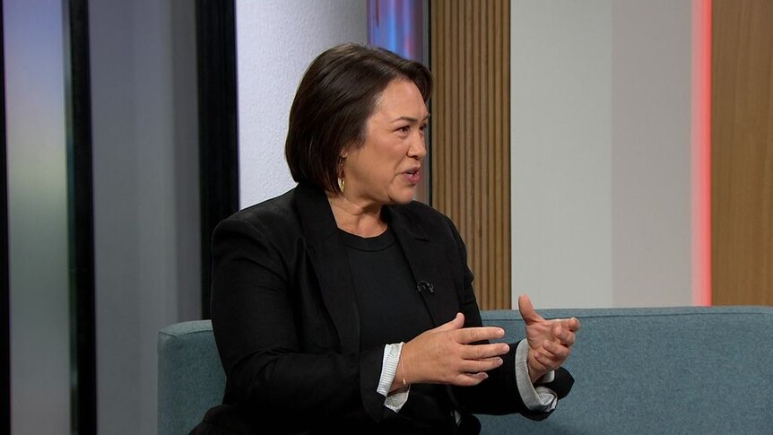 A woman sitting on a couch in a television studio speaks to someone off camera during an interview.