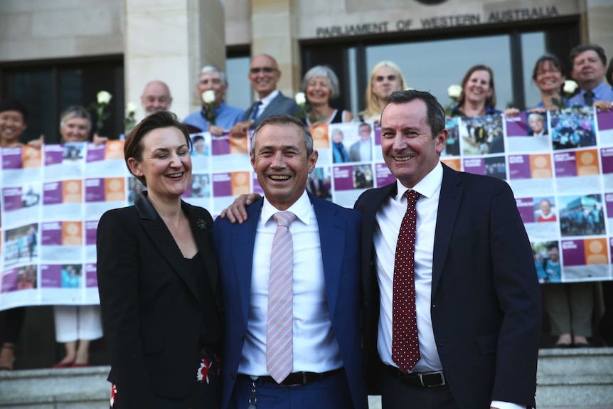 Three politicians on the steps of parliament with a group of pro-euthanasia supporters in the background holding roses
