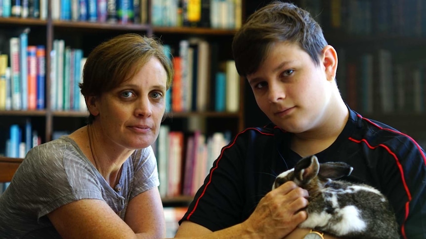 A boy holding a rabbit sits with his mother in front of a bookshelf