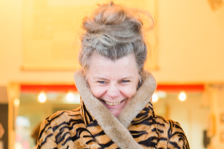A woman is standing in front of a bright background. She is smiling and hugging a big tiger-print coat around her.