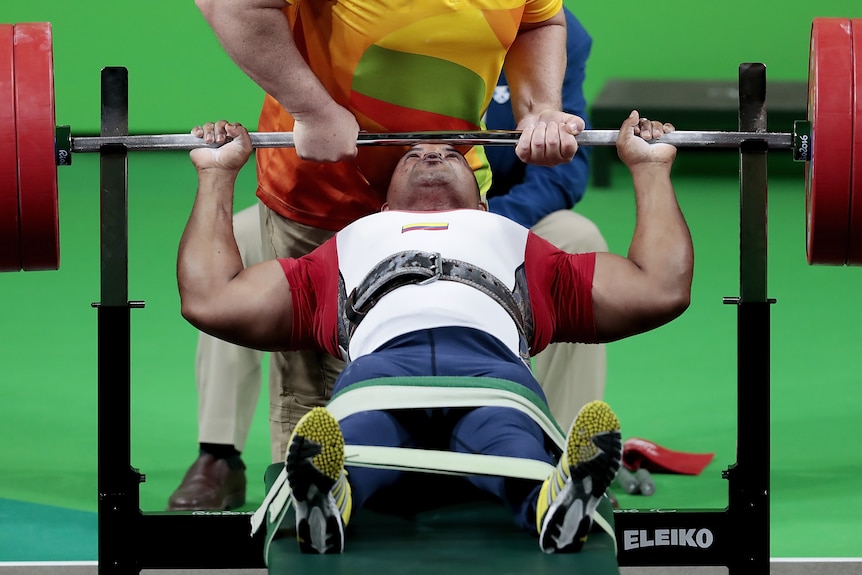 A Paralympic powerlifter grasps the bar while lying down with his legs strapped - a spotter also has the bar..