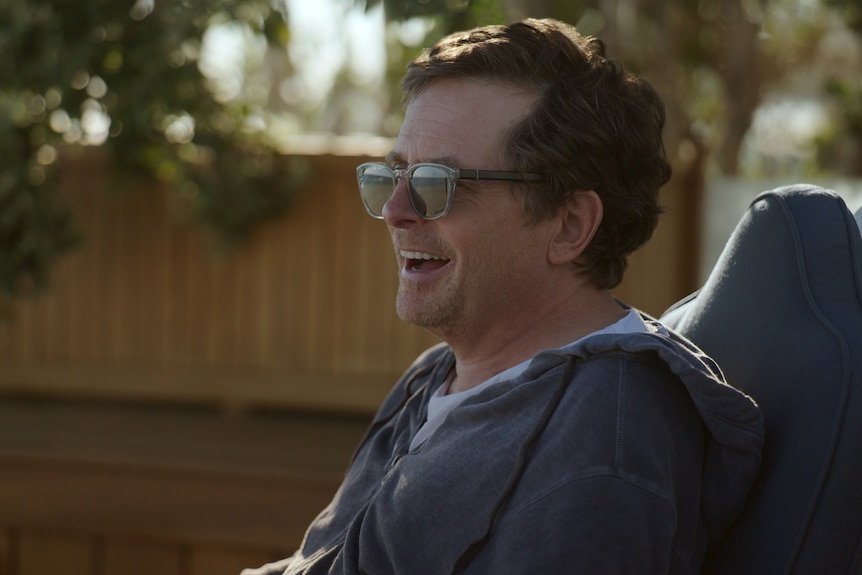 Actor Michael J. Fox, wearing sunglasses and a grey hoodie, smiles broadly, facing away from the camera.