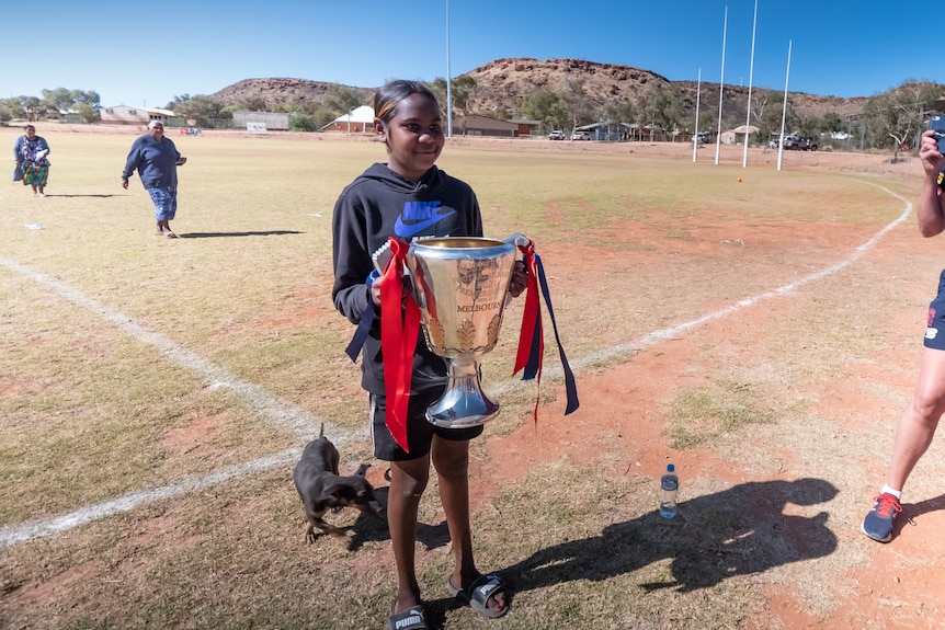 A child poses for a photo with the AFL premiership cup on a grassy oval with a rocky hill behind