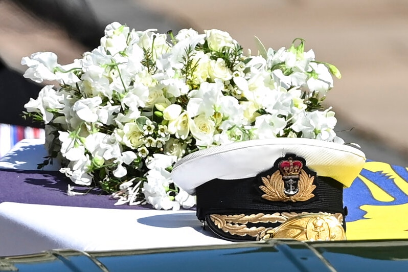 A Royal Navy cap sits on top of a coffin draped in a royal flag, with white lilies behind it.