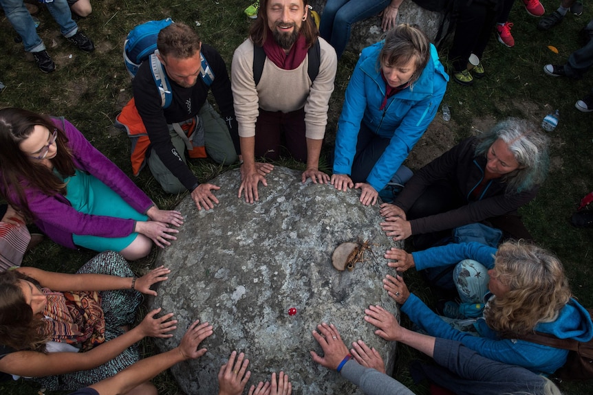 People with closed eyes, smiling and looking upwards place their hands on a large rock.