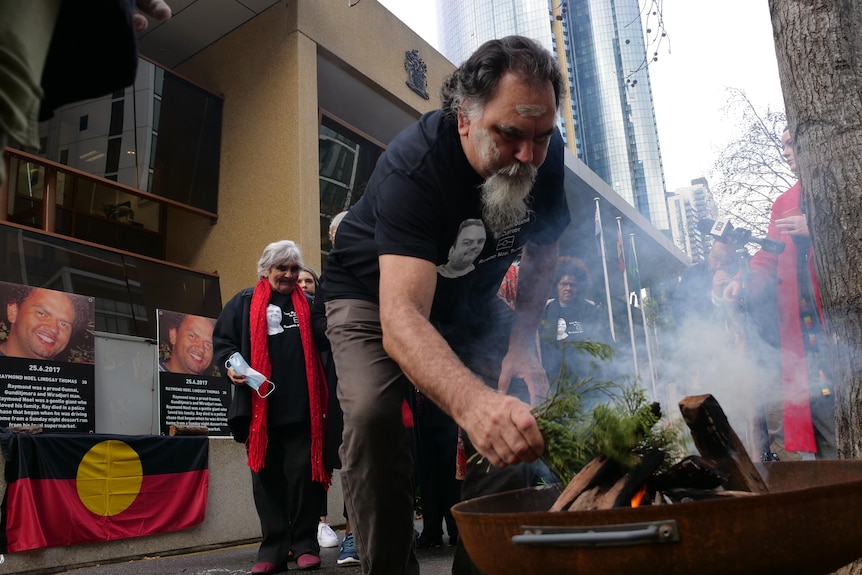 A man with a long beard places a plant into a fire during a smoking ceremony. 