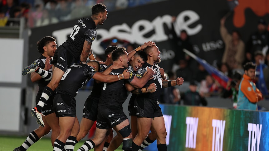 A New Zealand rugby player is surrounded by his teammates as he celebrates a try in front of the grandstand.