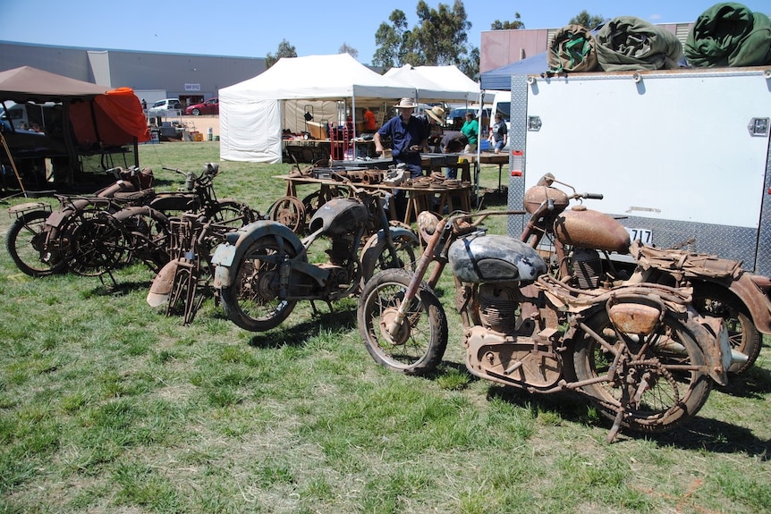 A bunch of rusty vintage motorcycles sit next to a trades stall
