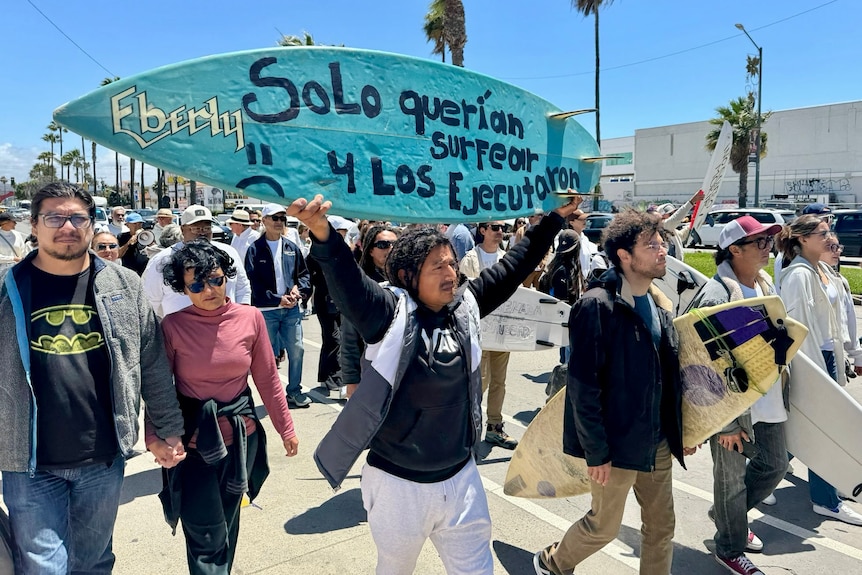 People march down a street surrounded by palm trees. Some hold surfboards with messages in Spanish.