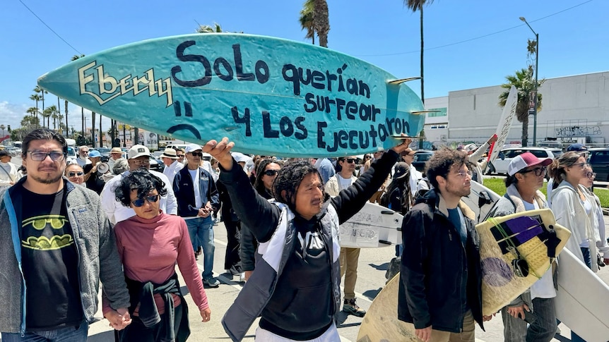 People march down a street surrounded by palm trees. Some hold surfboards with messages in Spanish.