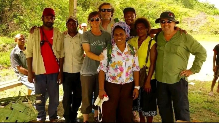 A group of people stand together smiling, the scene behind them is sunny and green