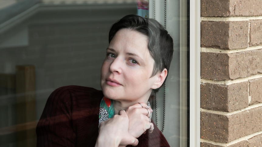 A white woman in her mid-30s with short brunette hair leans against a window, hands clasped