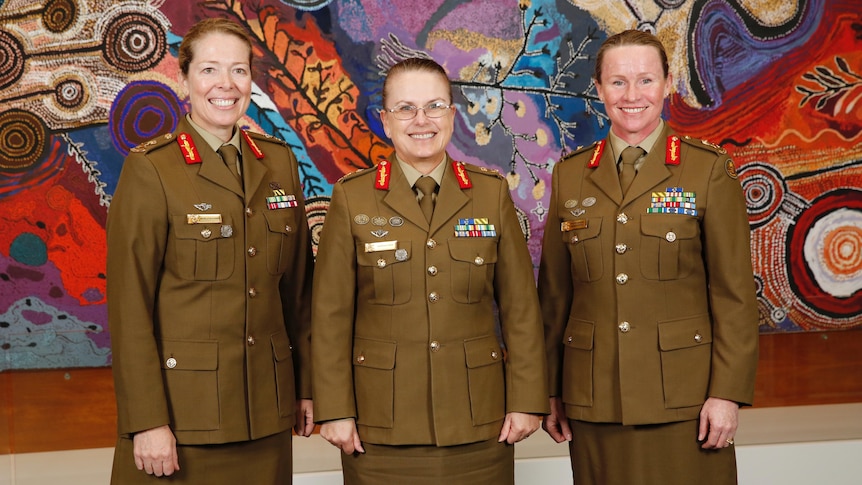 Three woman stand in army uniforms in front of a large and colourful aboriginal painting smiling for the camera