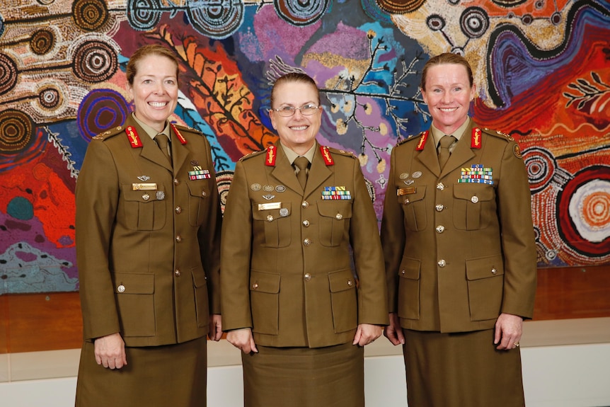Three woman stand in army uniforms in front of a large and colourful aboriginal painting smiling for the camera