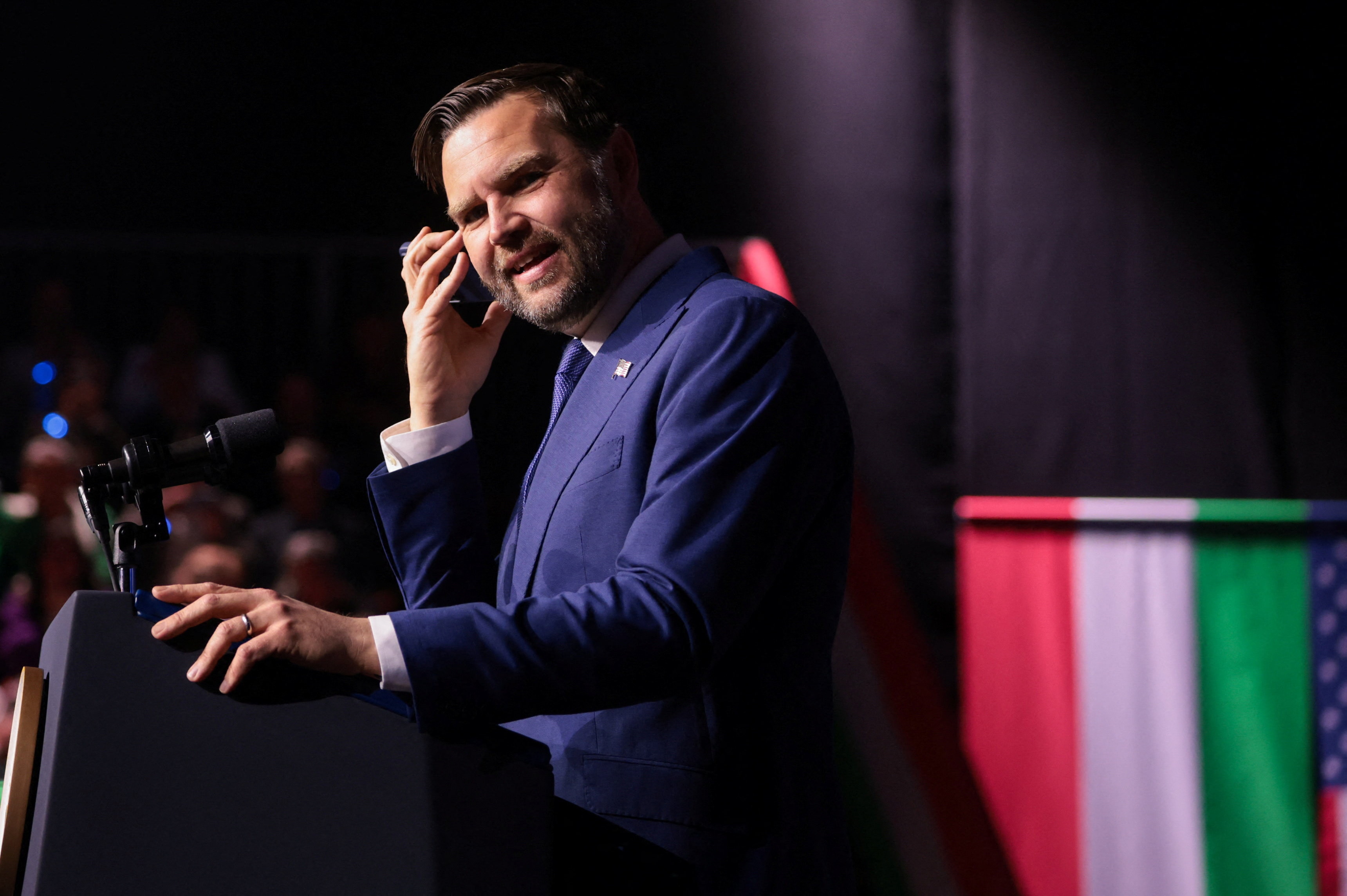 JD Vance smiles as he stands at a podium with a mobile phone to his ear.