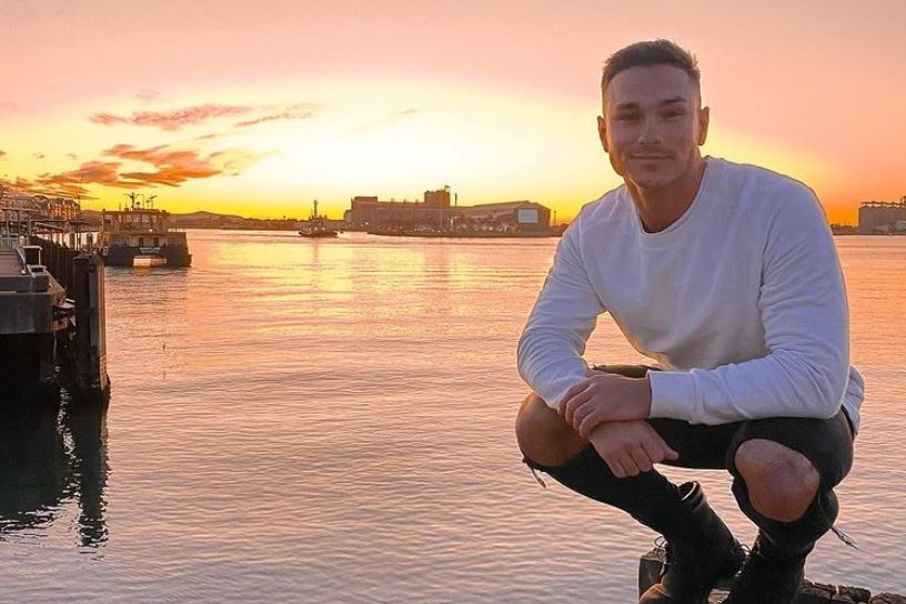 A smiling young man crouches on a pier in front of a waterway at sunset.