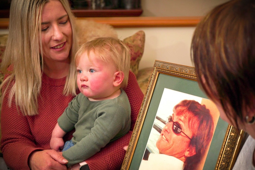 A baby boy poses next to a photo of a man. Two women are on either side of the baby, looking at him.