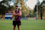 Older man wearing cap, singlet and shorts laughs while standing on country football oval