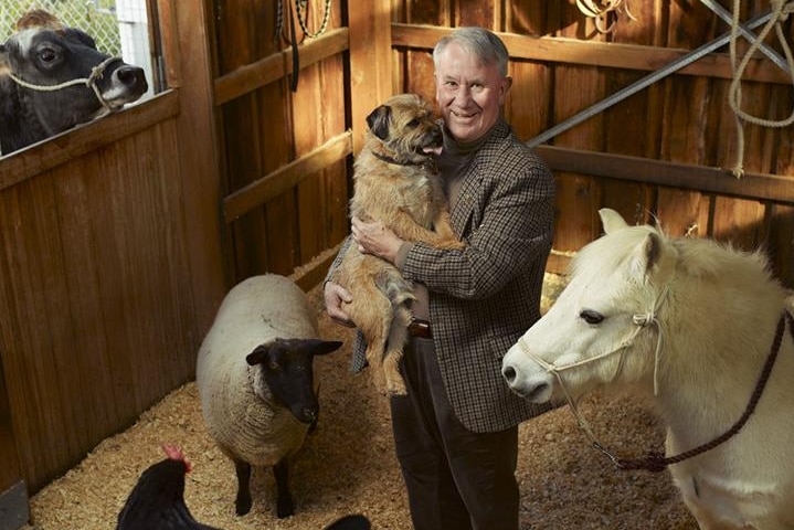 Dr Hugh Wirth stands in a barn surrounded by animals and holding a dog.