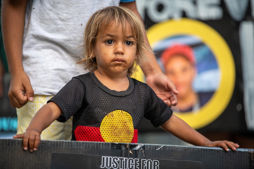 A young Indigenous boy wears a shirt with an Aboriginal flag pattern as he holds a sign calling for justice