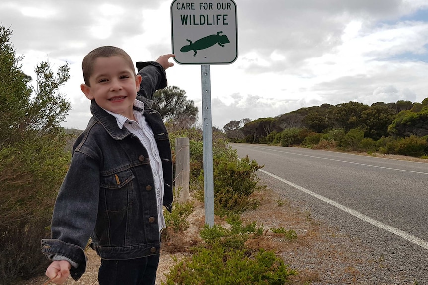 A young boy in denim jeans and a denim jacket smiles as he points to a sign that says, "Care for our wildlife".