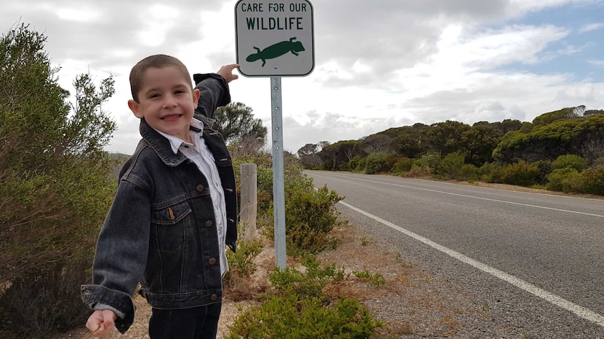 A young boy in denim jeans and a denim jacket smiles as he points to a sign that says, "Care for our wildlife".