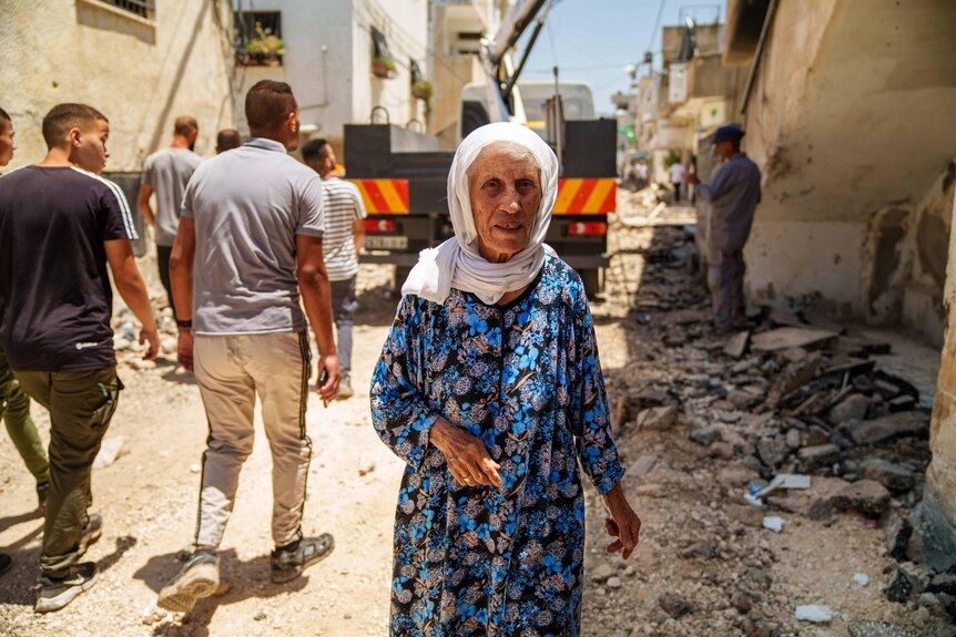 An older woman in a veil walks through rubble