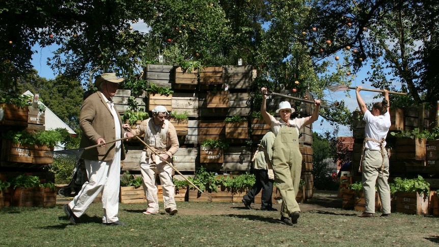 Five performers in overalls carrying gardening tools on a grass stage in front of stacked boxes of plants.