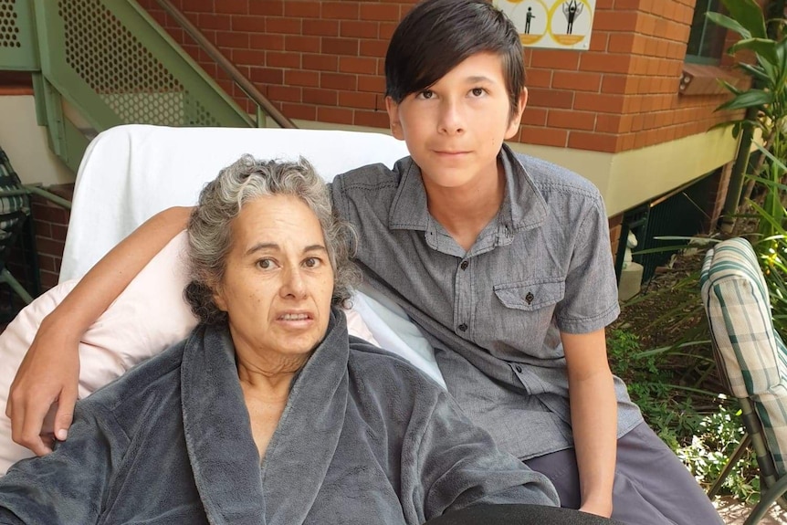 a young boy sits on a bed where his mother lays, both of them are looking at the camera