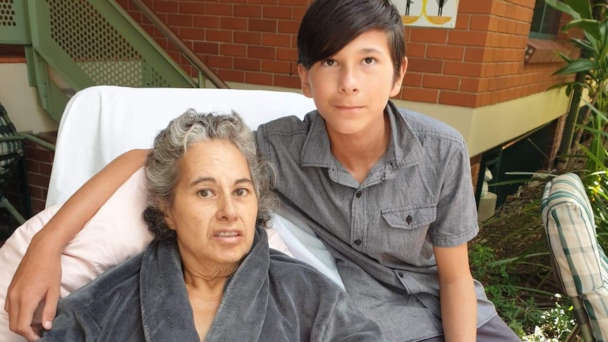 a young boy sits on a bed where his mother lays, both of them are looking at the camera