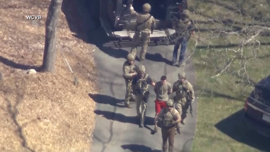 Jack Teixeira, in T-shirt and shorts, being taken into custody by armed tactical agents.