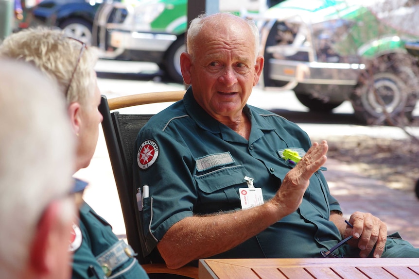 Volunteer ambulance officer in uniform sits at table talking to others, not identifiable