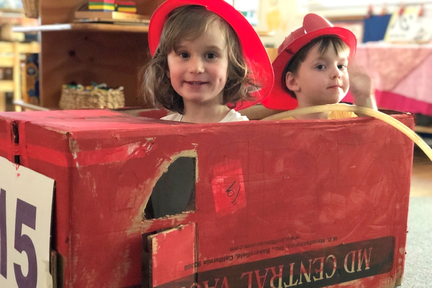 Two kindergarten boys wearing firefighter dress ups in a makeshift red, cardboard fire truck.
