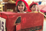 Two kindergarten boys wearing firefighter dress ups in a makeshift red, cardboard fire truck.