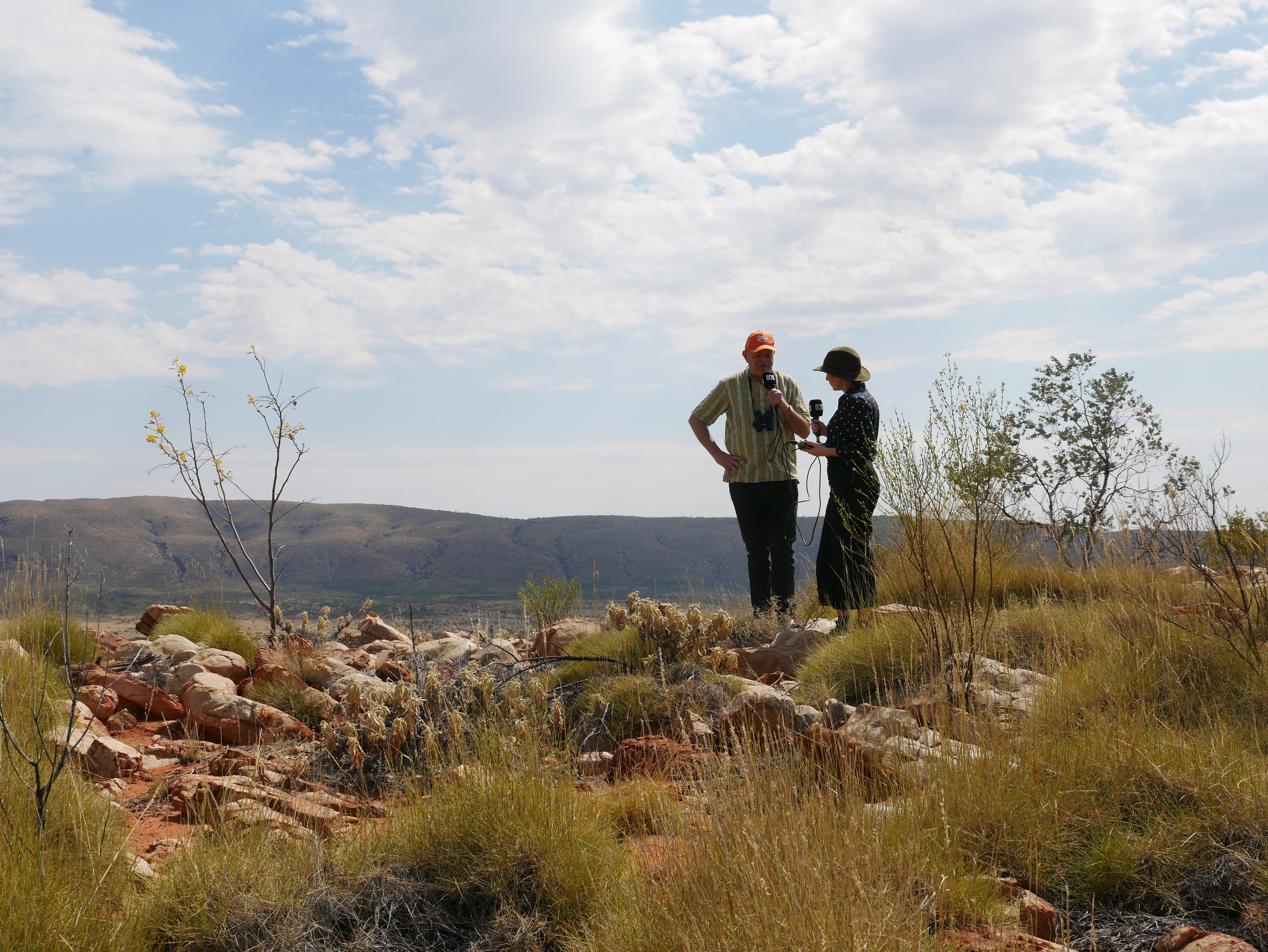 A man and a woman stand atop a mountain in the outback, talking. 