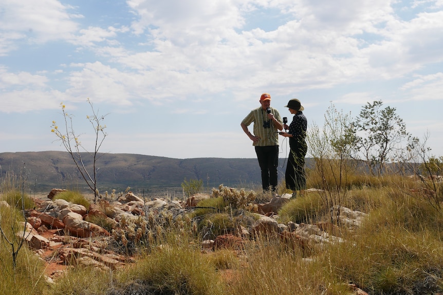 A man and a woman stand atop a mountain in the outback, talking. 