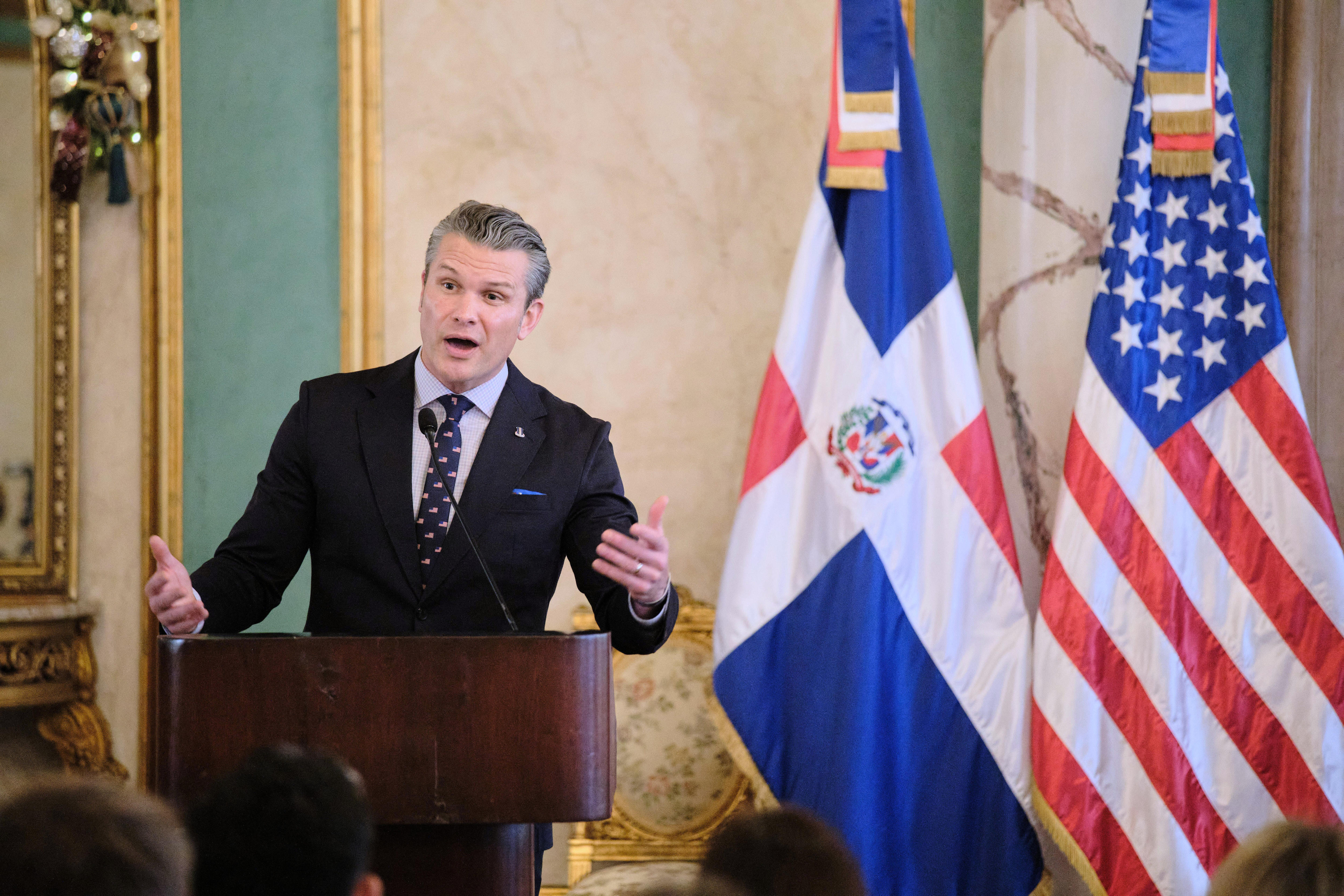 Pete Hegseth in a black suit speaking at a dark wood lectern and gesturing with both hands, next to a US and Dominican flag