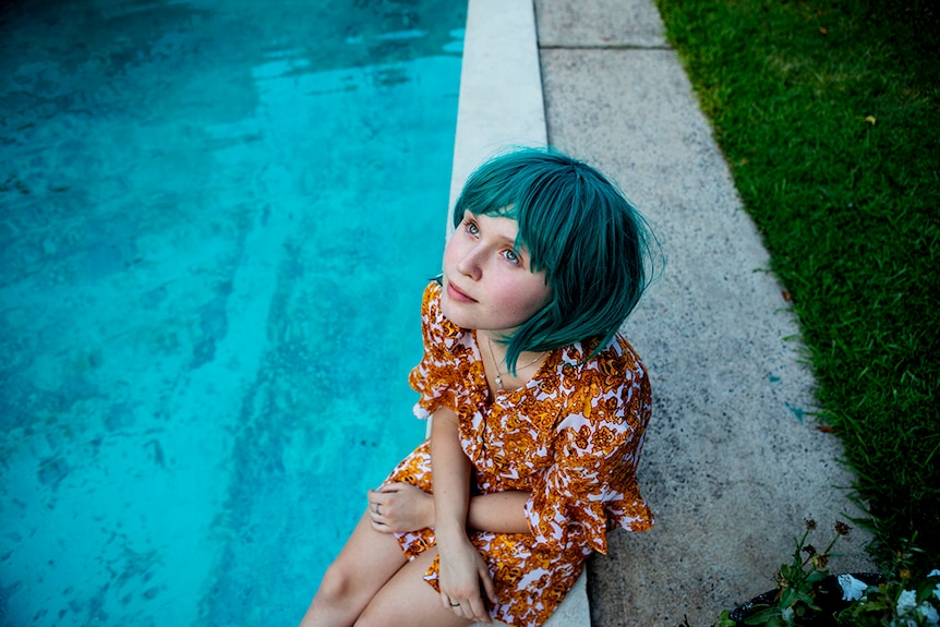 A young girl wears cyan bob wig and orange and white pattern outfit and sits on concrete edge of swimming pool and looks to sky.