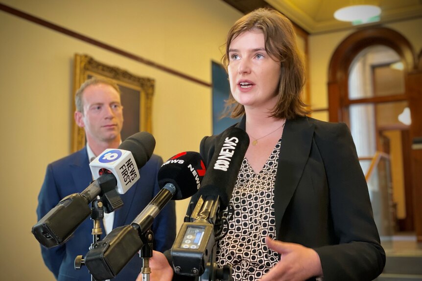 A woman stands in front of media microphones talking while a man stands in the background looking at her