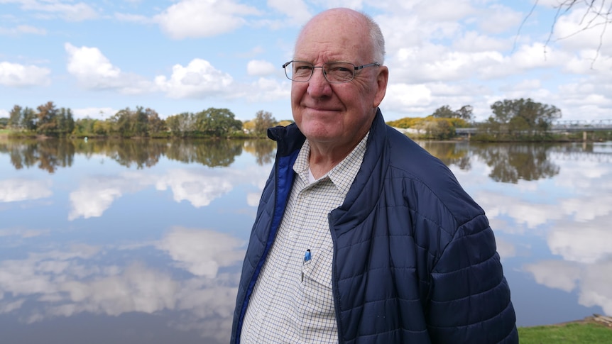 An older man wearing a shirt and blue jacket stands in front of river in a regional area.