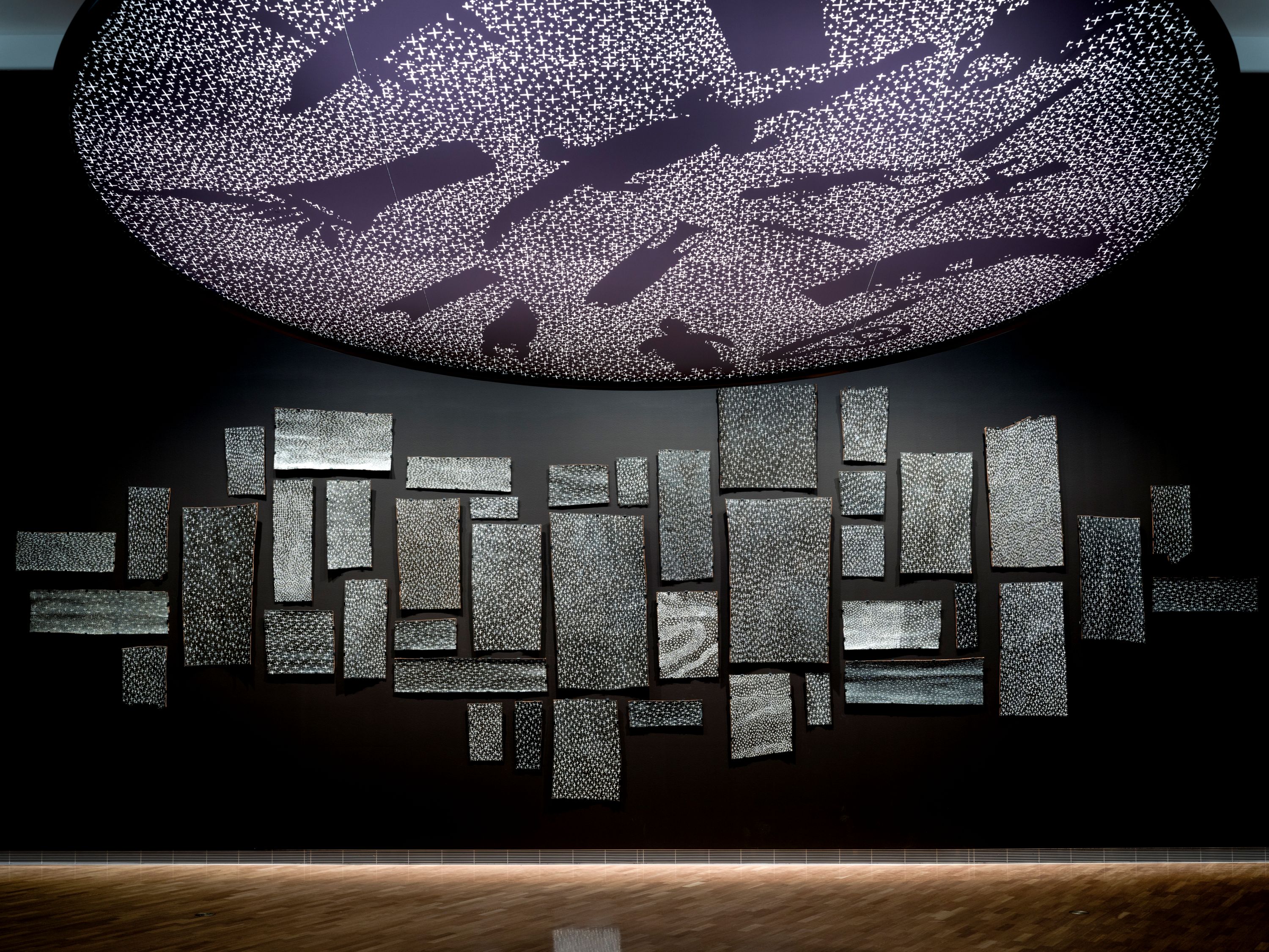 En una galería, un disco violeta cuelga del techo, iluminado con formas de animales. En las paredes cuelgan cuadros de corteza con estrellas.