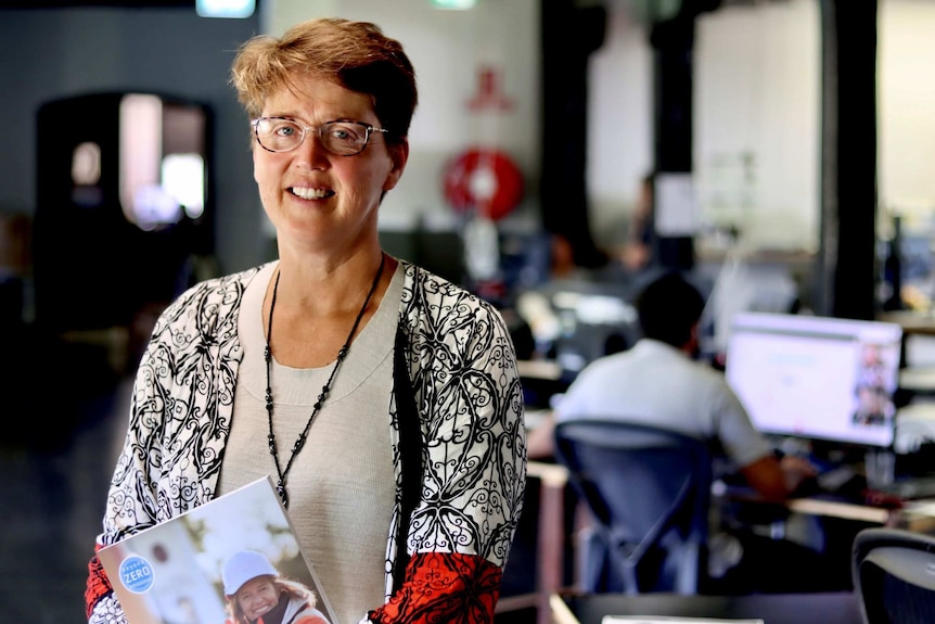 A women with short, light red hair stands in an office, holding a coloured CSIRO document 