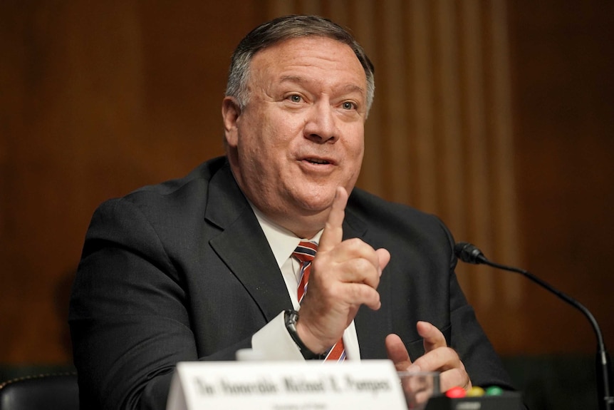 A man in a suit raises his right index finger for emphasis as he speaks into a microphone that has been mounted to a desk.