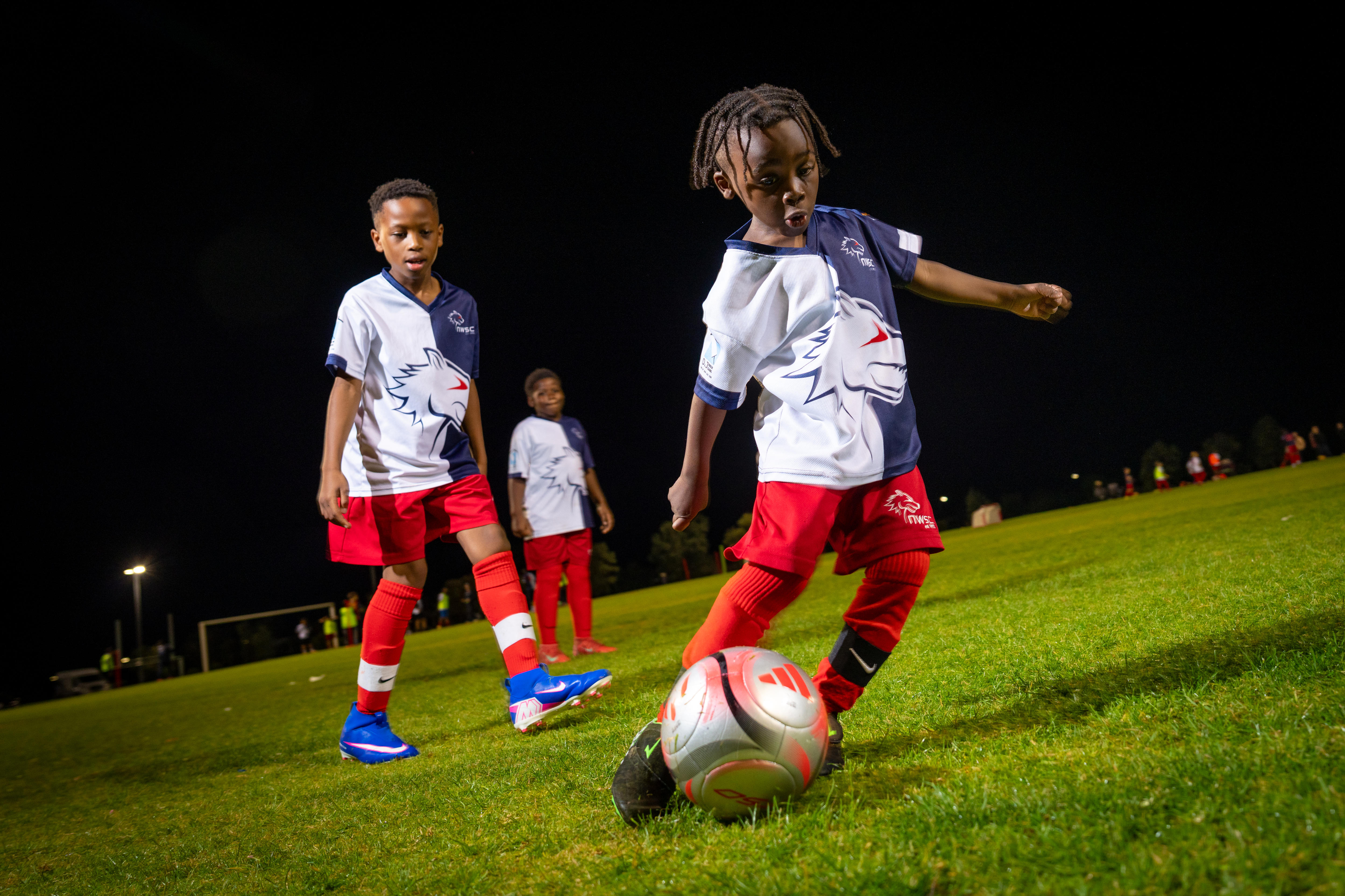 Three young boys in soccer uniform, one of whom is kicking a ball